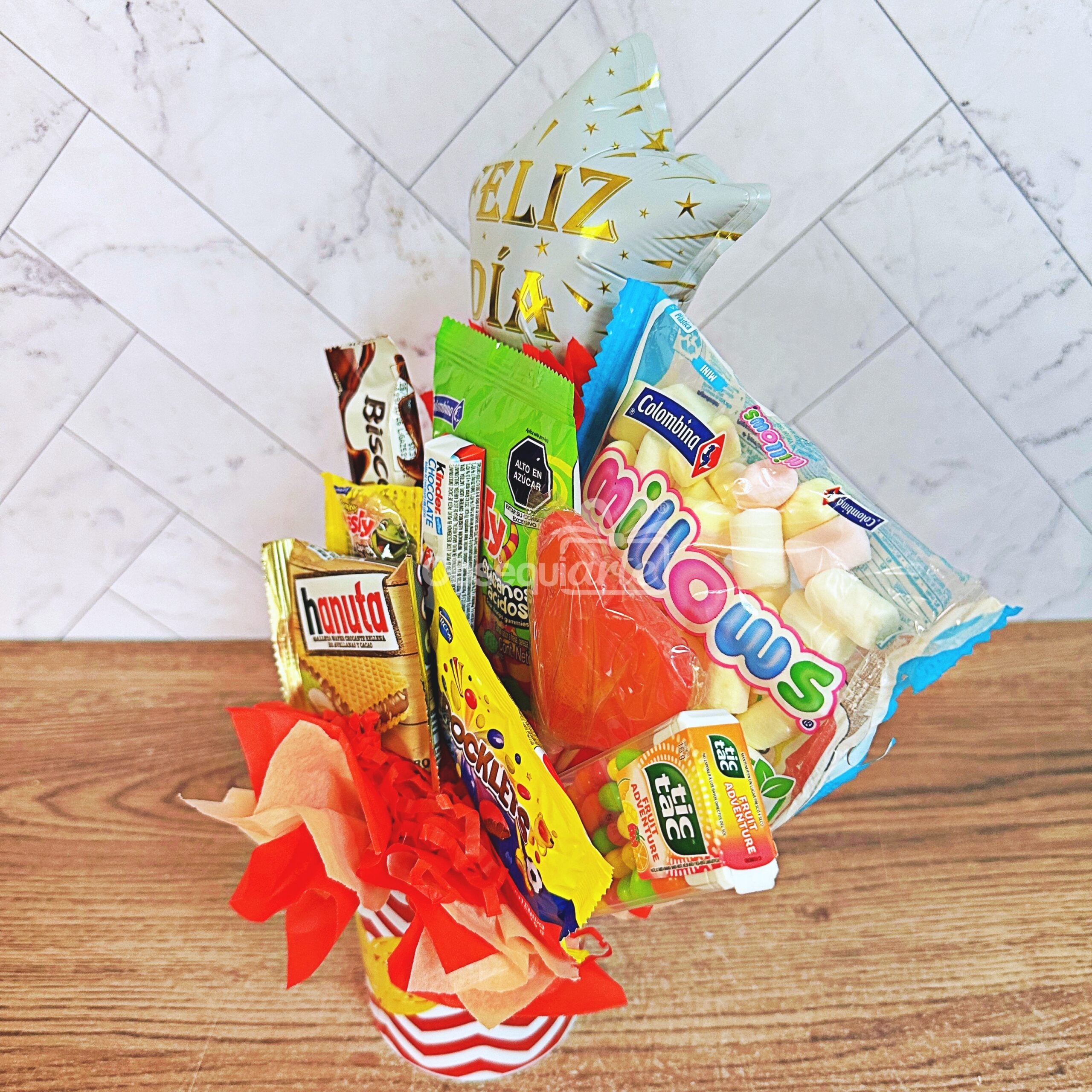 El Regalo Jarro Mi Corazón incluye una selección de aperitivos y dulces dispuesta a modo de ramo, complementada con un globo en forma de estrella con la inscripción «Feliz día», todo ello presentado de forma elegante en un recipiente a rayas con la etiqueta «Mi Corazón» y colocado sobre una base de madera, lo que lo convierte en el regalo perfecto para alegrarle el día a cualquiera.
