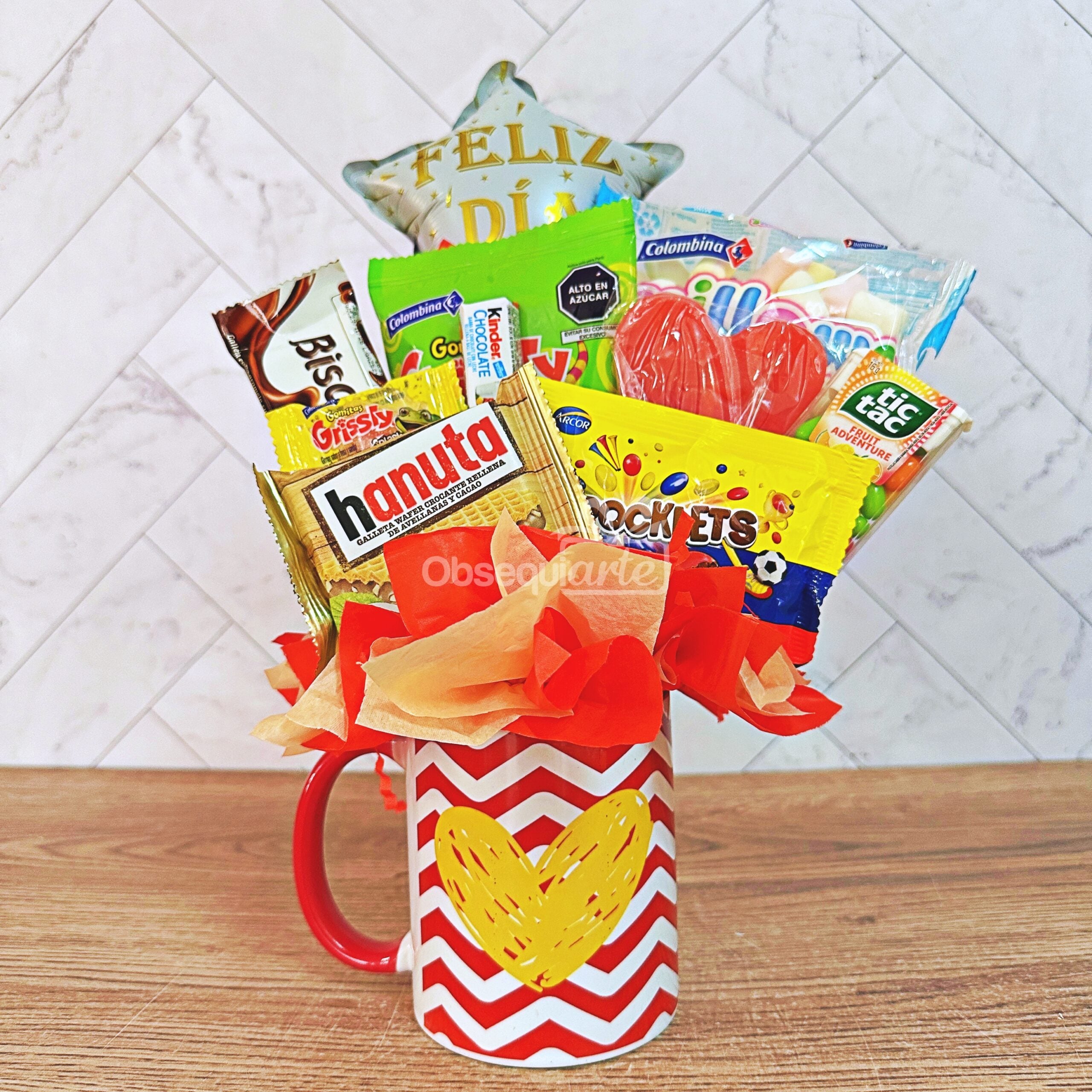 El «Regalo Jarro Mi Corazón» consiste en una taza roja y blanca decorada con un corazón amarillo y la inscripción «Mi Corazón», llena de aperitivos y dulces variados, y coronada con un globo en forma de estrella con la inscripción «Feliz Día», todo ello dispuesto artísticamente a modo de ramo de regalo sobre una superficie de madera.