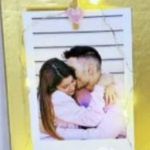 Una foto de una pareja sentada muy cerca y abrazándose, sujeta a un tablero dorado con cuerdas de luces alrededor de la imagen.