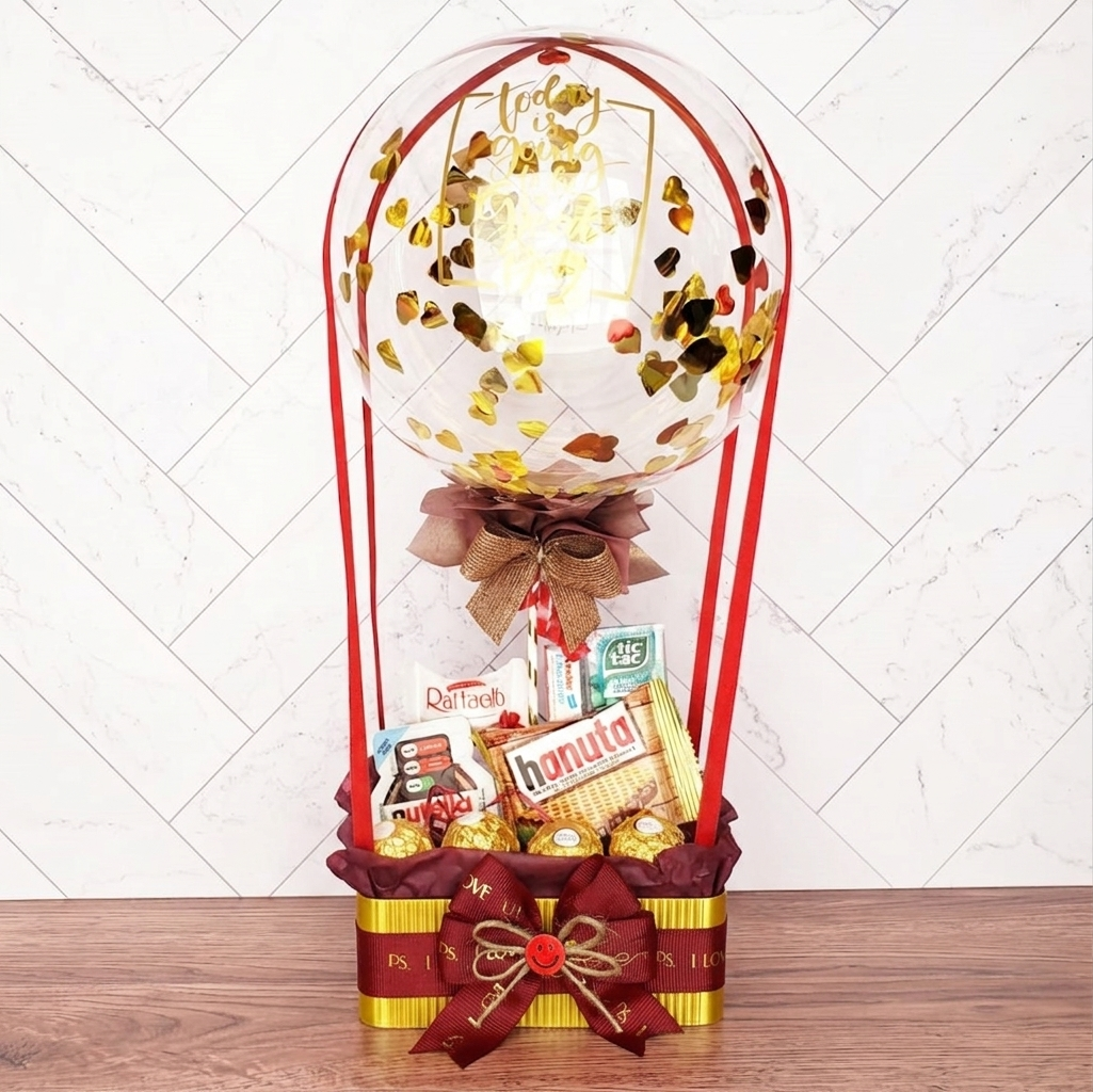 Regalo Regalo Gran Cesta Ferrero con globo, cintas rojas y lazo, incluye barquillos Hanuta, bombones y aperitivos variados expuestos sobre una superficie de madera.