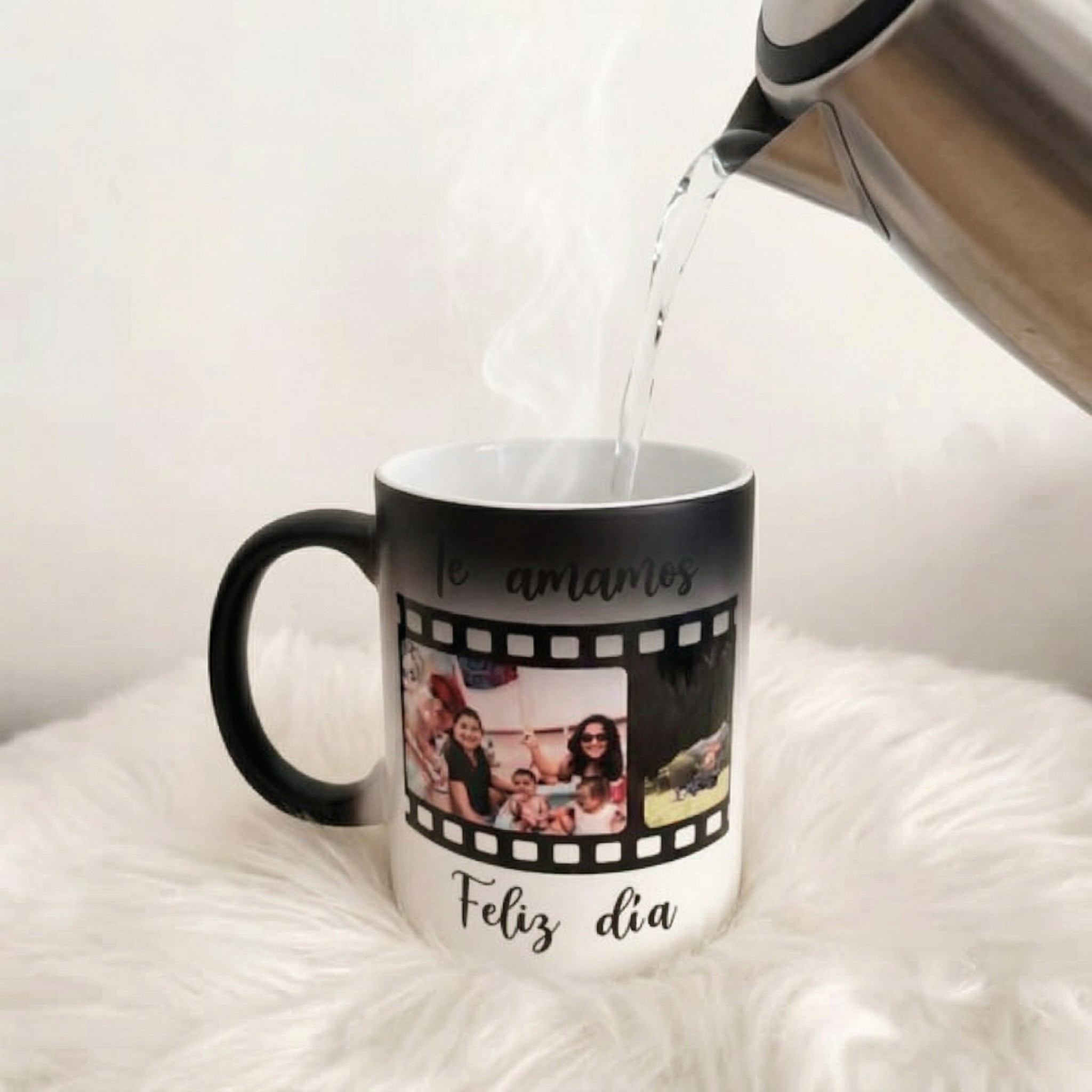 Una tetera vierte agua caliente en un Regalo Taza Mágica con fotos familiares y el texto en español "Te amamos, Feliz día", mientras la taza descansa sobre una superficie blanca y peluda.