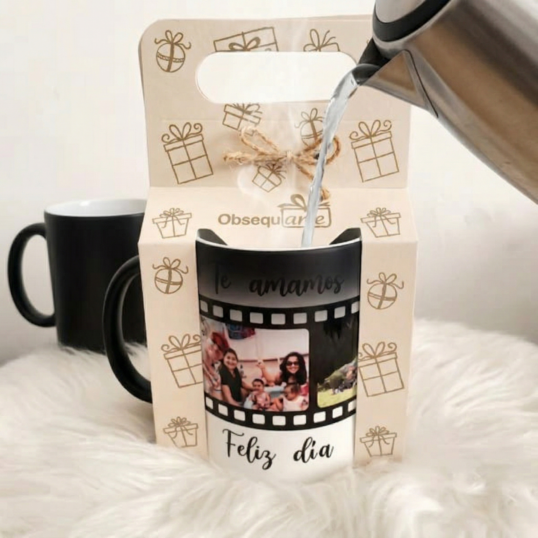 Se vierte una bebida caliente en un Regalo Taza Mágica con fotos familiares y las palabras "Te amamos" y "Feliz día", colocado delante de una caja de regalo, con una taza negra visible al fondo.