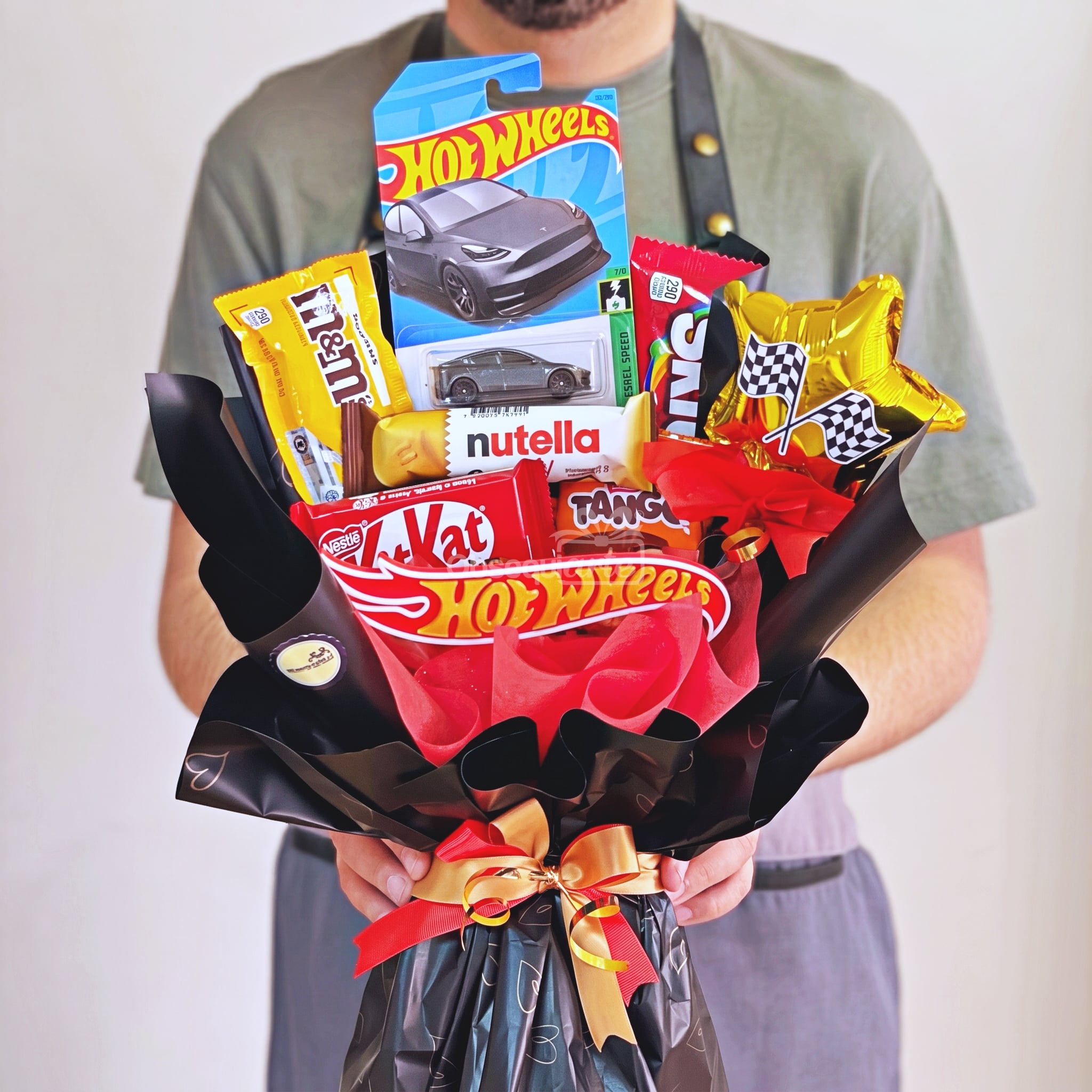 Una persona sostiene un «Ramo Hot Wheels» —un ramo que incluye aperitivos variados, un cochecito de juguete Hot Wheels en miniatura y dos globos con estrellas doradas—, envuelto con elegancia en papel negro y rojo con una cinta.