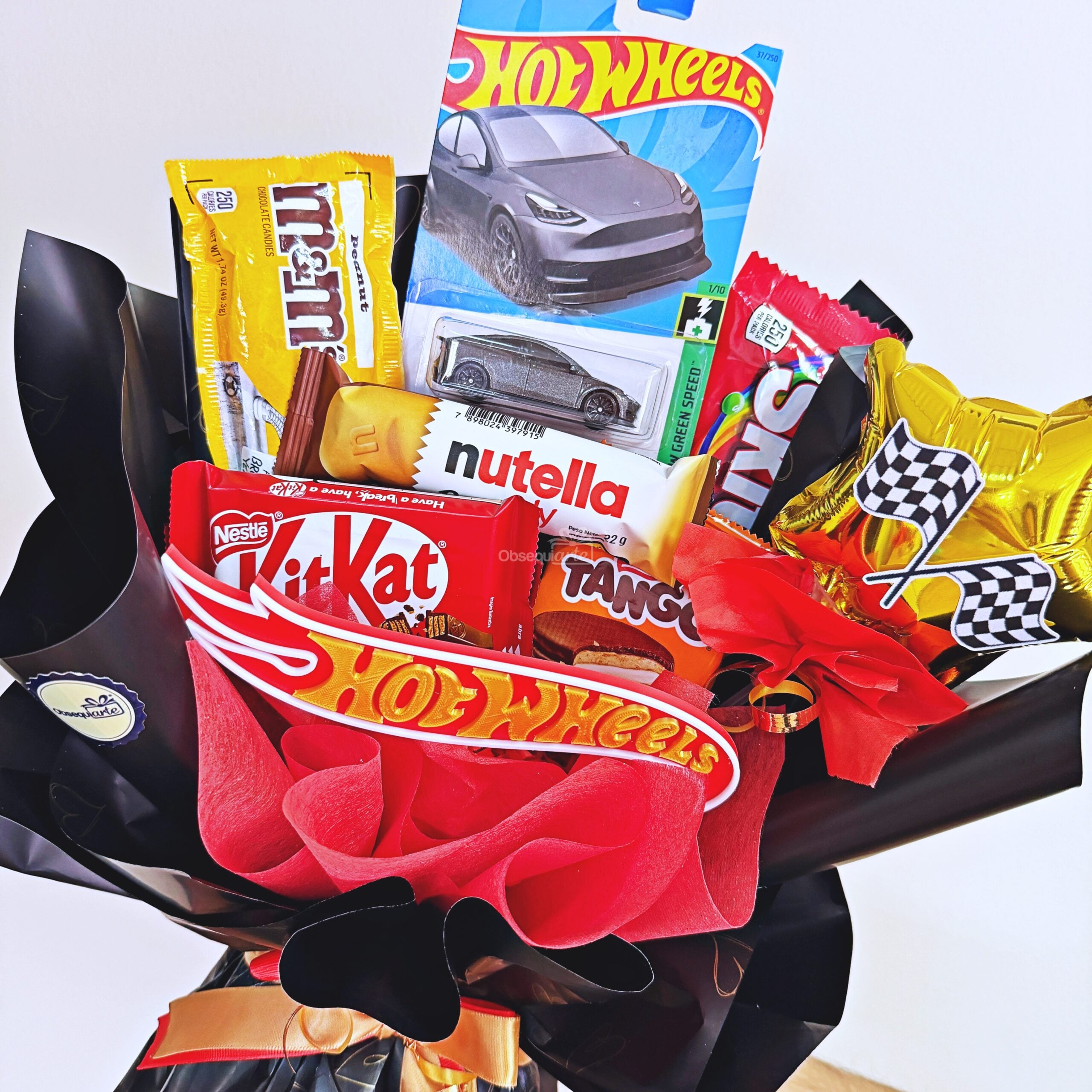 Te presentamos «Ramo Hot Wheels»: un arreglo único en su género que combina coches de juguete Hot Wheels, Kit Kat, M&M’s, Nutella, Tango y globos con estrellas doradas, todo ello envuelto con elegancia en un llamativo papel negro y rojo.