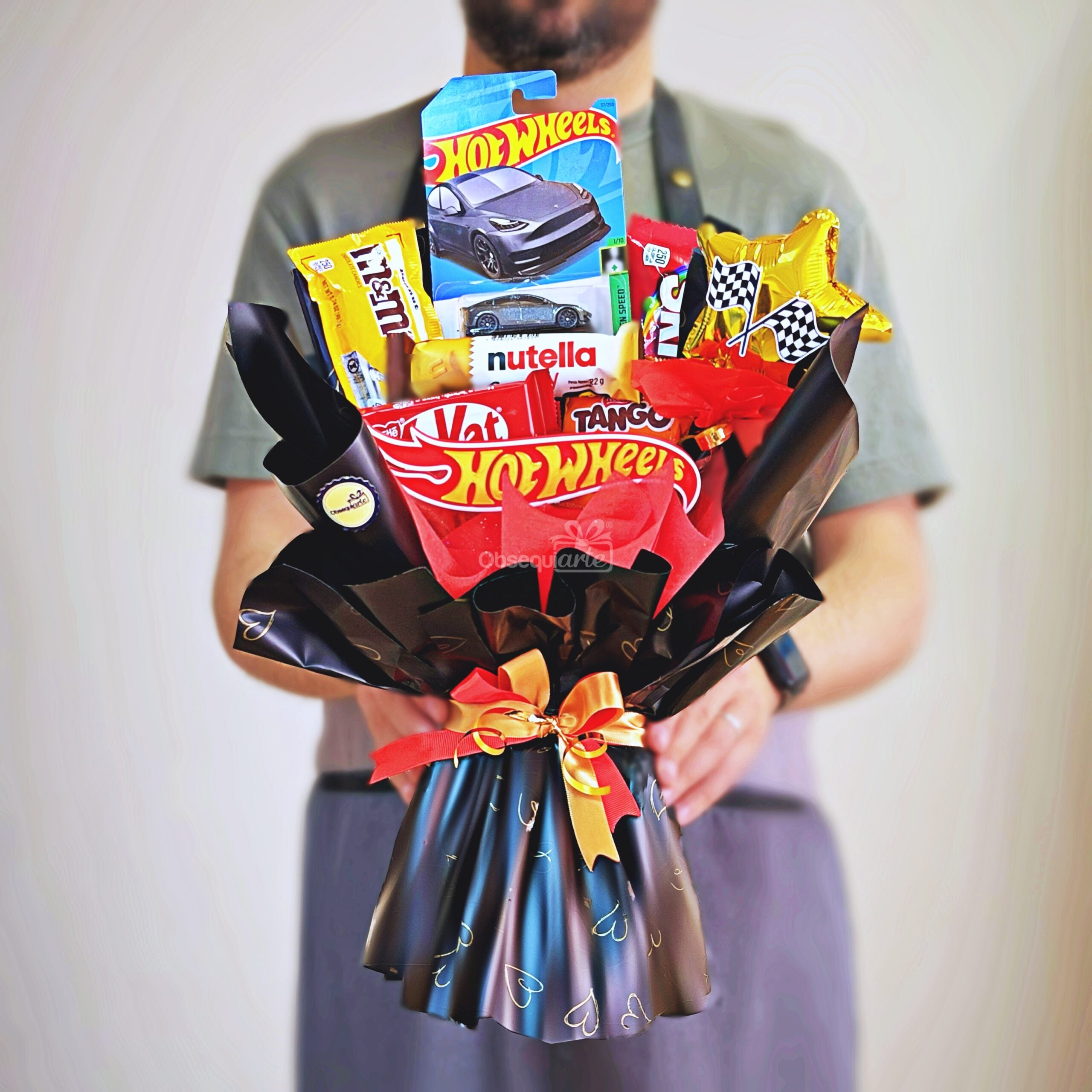 Una persona sostiene un llamativo ramo «Ramo Hot Wheels», que incluye tabletas de chocolate, un tarro pequeño de Nutella, un cochecito de juguete Hot Wheels y un envoltorio decorativo atado con una cinta roja.