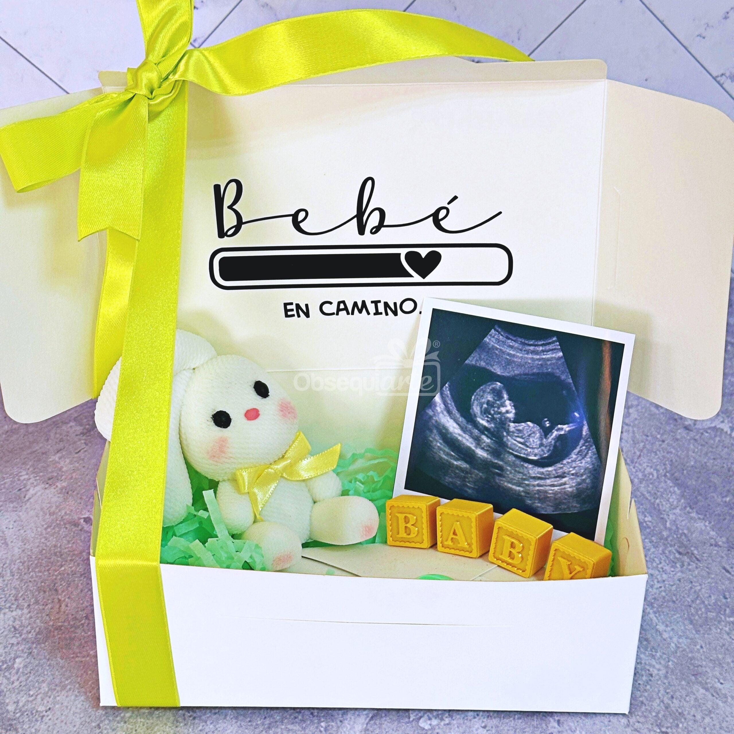 El «Regalo Bebé en Camino» incluye una caja abierta adornada con un lazo amarillo, un conejito de peluche muy tierno, bloques de juguete para bebés que forman la palabra «BABY» y una ecografía, lo que lo convierte en el regalo perfecto para un bebé. La tapa está decorada con la frase «Bebé en camino» junto a un precioso dibujo de un corazón.